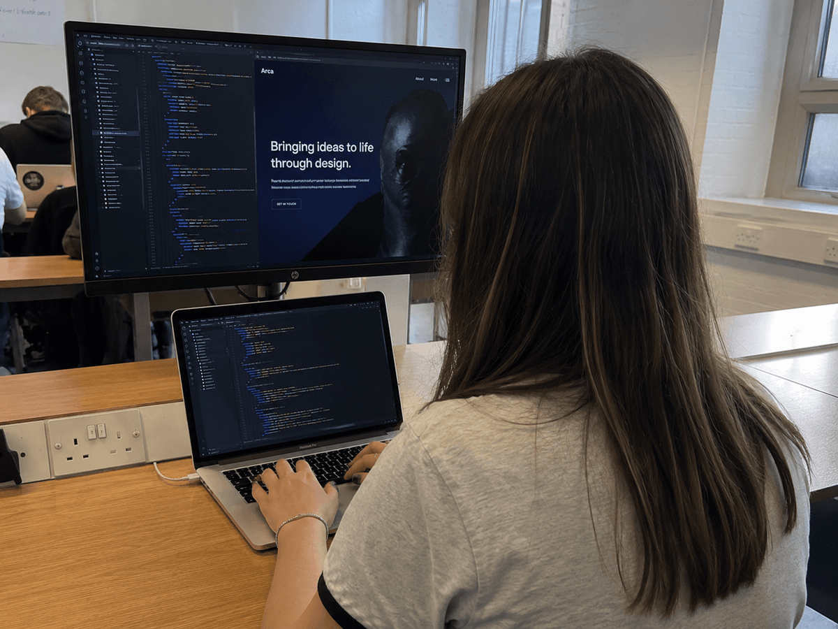 A student at a laptop and monitor, with code on one screen and a website design on the other